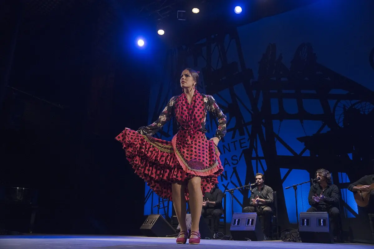 A flamenco dancer in a red polka-dot dress stands with a hand on her skirt on a stage lit in blue, with singers and guitarists seated behind her