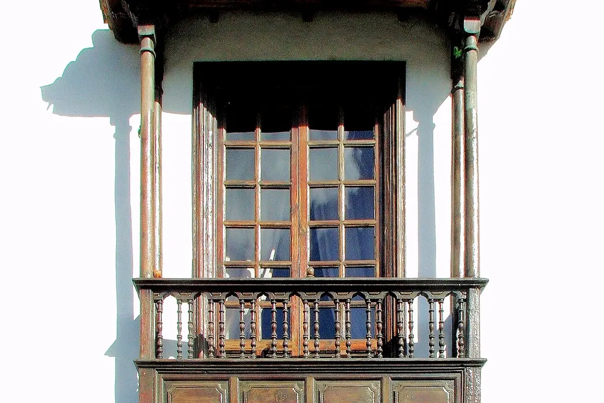A wooden balcony with a tiled roof above a whitewashed wall with a stone corner, seen from below