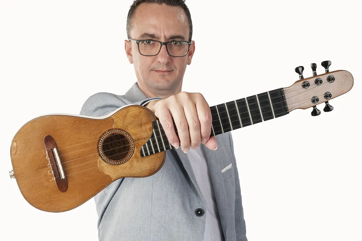 A man in glasses and a grey jacket holds a timple, a small five-string Canarian instrument with a pale headstock, towards the camera